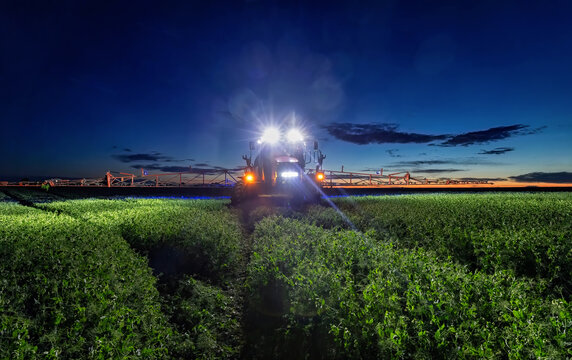 Agricultural self-propelled sprayer working in a green field at dusk with illuminated headlights.