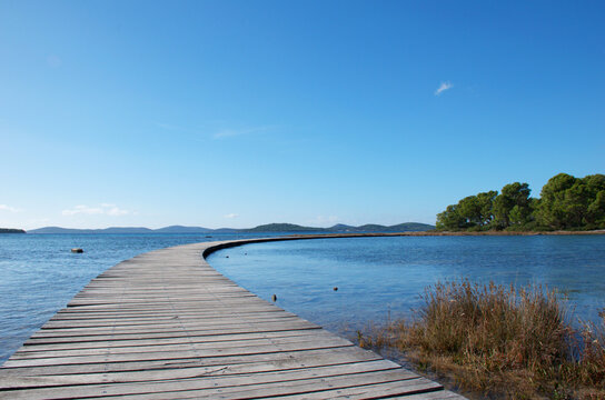 A curved wooden boardwalk leads into the calm, blue sea beneath a clear summer sky. The winding path evokes a sense of travel, freedom, Zen, purpose, possibility, and peaceful retreat. 