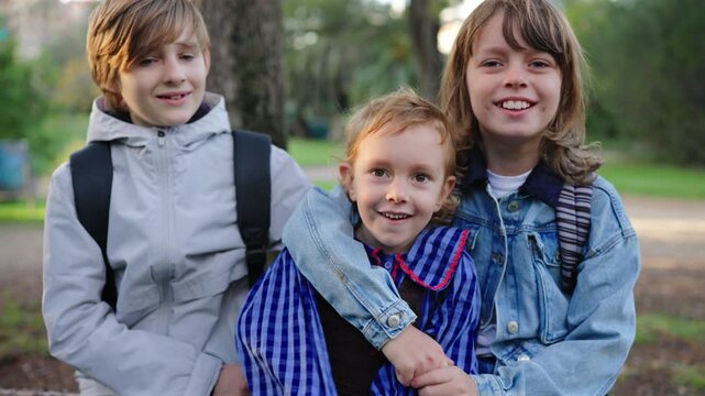 Three children siblings pose together on a fallen tree log in a lush green park. Older boy in gray jacket and girl in denim hug their younger sibling wearing striped blue shirt.