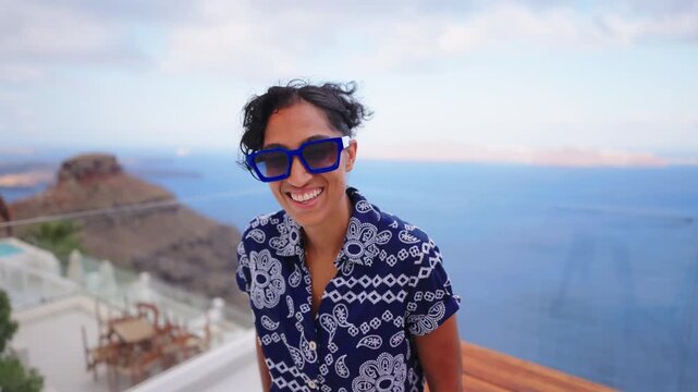 Happy young woman wearing stylish blue sunglasses and patterned shirt smiles at camera on white terrace overlooking Aegean Sea in Santorini Greece with volcanic cliffs in the background at dusk.