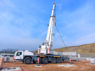 Heavy-duty white mobile crane lifting a long precast concrete beam at a dirt construction site under a clear blue sky. © Framelum