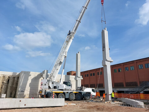 White mobile crane lifting a heavy precast concrete pillar while two construction workers guide it into place at a building site.