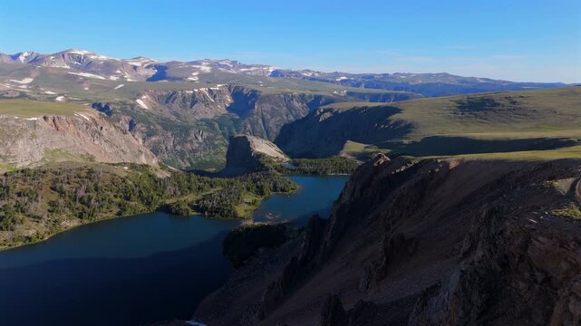 Aerial drone footage of Beartooth Highway Montana showcases stunning twin alpine lakes nestled among rugged mountain cliffs with snow-capped peaks, lush green meadows.