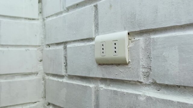 Hand plugging orange cable into wall electrical outlet, brick wall.