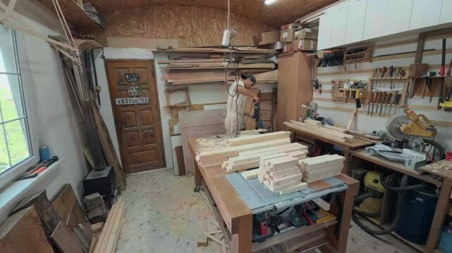 Dedicated self employed craftsman operating heavy drill machine to cut plywood circles inside cluttered private home workshop during wide angle shaky handheld documentary walking observer POV shot