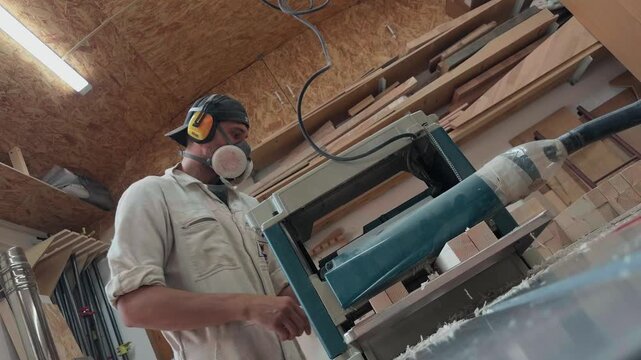 Focused male craftsman wearing protective respirator and industrial earmuffs feeding timber blanks into thickness planer machine on wooden workbench in authentic bright carpentry workshop low angle