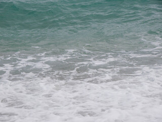 Fototapeta premium Close-up of turquoise-green Adriatic wave breaking with white foam patterns on Budva beach, Montenegro. Translucent wave crest, foamy wash below. Vivid abstract seascape with colour and texture.