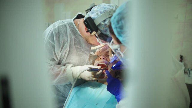  A dentists drilling a hole for a dental implant in the jaw of a young adult female patient with a dental drill, while draining excess saliva. Footage of a real-life surgical procedure