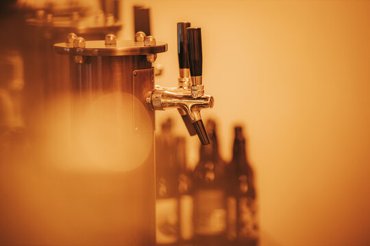 Beer tap with black handles and shiny chrome finish, positioned against a blurred background of dark beer bottles in a warm-toned bar setting
