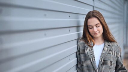 Fototapeta premium Serene businesswoman in checkered blazer enjoys moment of peace, leaning against grey metal wall with closed eyes