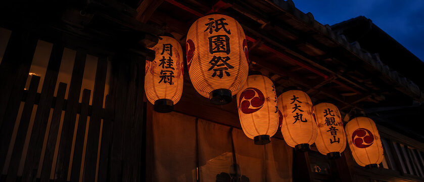 Dramatic low-angle shot of glowing Japanese chochin paper lanterns hanging from dark wooden machiya during Gion Matsuri night &mdash; Kyoto festival atmosphere