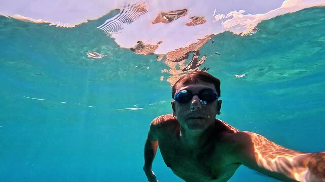 A man swims underwater in the Mediterranean Sea. The man is of Caucasian origin, is forty-two years old, has short hair and has swimming goggles on his face