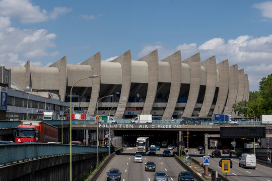 Exterior view of the Parc des Princes stadium in Paris with heavy traffic on the Boulevard Peripherique highway passing underneath the iconic brutalist concrete architecture on a sunny day