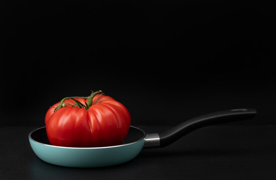 There is a red tomato in a pan. The pan and the tomato are set against a dark background. There is space for text.