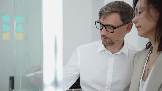 Business professionals collaborating and brainstorming ideas on a glass wall with sticky notes, organizing tasks and developing a project strategy in a modern office meeting