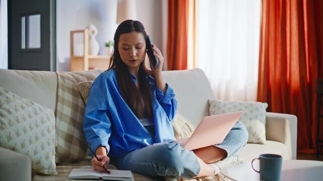 Asian woman talking phone working laptop sofa. Freelancer taking notes at home