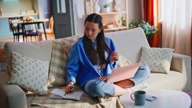 Asian student writing notes working pink laptop sofa. Woman studying at home.