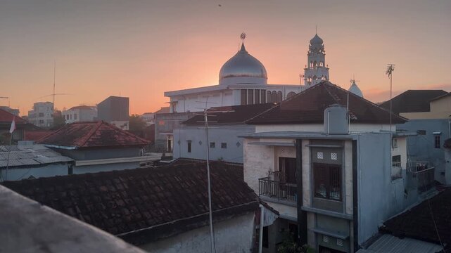 Timelapse or real time footage of a white mosque with dome and minaret during golden sunrise. View of traditional Indonesian residential rooftops in the foreground with warm orange sky