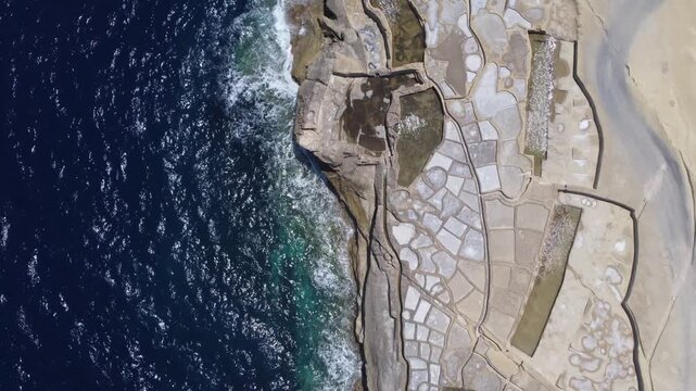Xwejni s centuries-old salt pans unfold along the rugged coast of Zebbug, Gozo, geometric pools and vivid Mediterranean blues in a sweeping summer Aerial view. High quality 4k footage