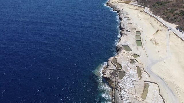 Xwejni s centuries-old salt pans unfold along the rugged coast of Zebbug, Gozo, geometric pools and vivid Mediterranean blues in a sweeping summer Aerial view. High quality 4k footage
