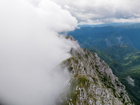 Aerial view of Piatra Craiului Mountains with rocky ridges partially obscured by thick clouds and lush green slopes in Zarnesti, Brasov, Romania.
