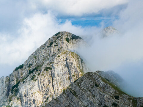 Aerial view of Piatra Craiului Mountains featuring jagged limestone peaks partially obscured by thick white clouds and patches of green vegetation in Zarnesti, Brasov, Romania.