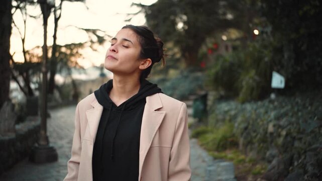 Young Hispanic woman walking through an urban park, stopping to breathe deeply and enjoy the fresh air, promoting mindfulness, wellness, balance and a healthy outdoor lifestyle.

