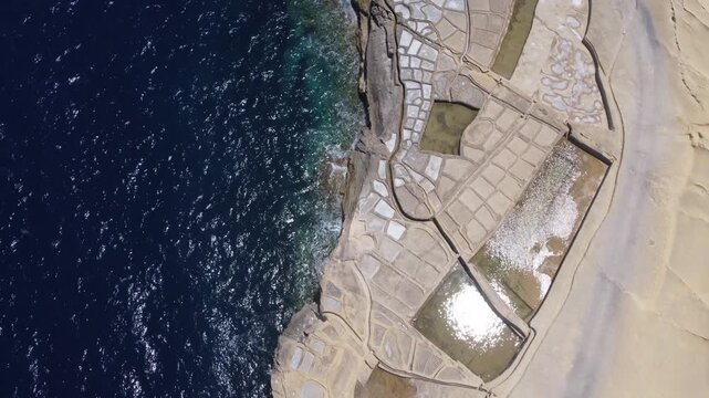 Xwejni s centuries-old salt pans unfold along the rugged coast of Zebbug, Gozo, geometric pools and vivid Mediterranean blues in a sweeping summer Aerial view. High quality 4k footage