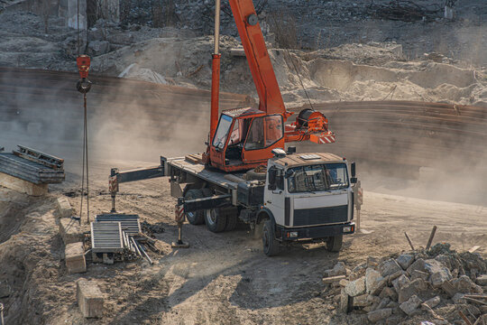 Construction scene featuring mobile crane mounted on truck, actively lifting heavy load, kicking up dust and debris. Truck with flatbed trailer equipped for heavy-duty work. Uneven ground with piles