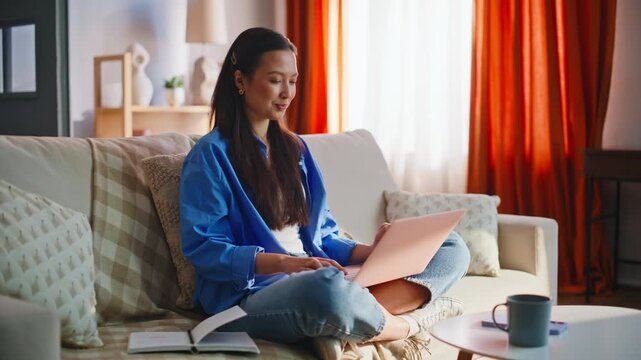 Asian woman working laptop home sofa. Freelancer typing notes living room.