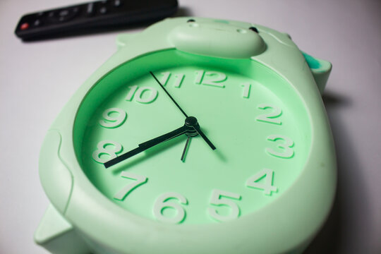 Close-up of a cute pastel green animal-shaped analog alarm clock on a white surface with a blurred remote control in the background.