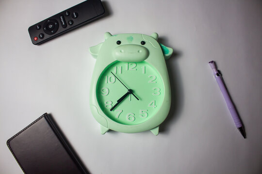 Flat lay of a cute pastel green cow-shaped alarm clock on a white workspace desk with a notebook, pen, and remote control
