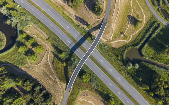 Aerial view of a bicycle and pedestrian bridge crossing a highway, surrounded by lush green fields, trees, and small ponds in Heerhugowaard, Noord-Holland, Netherlands.