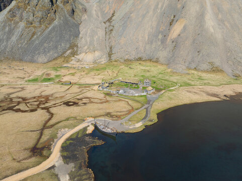 Aerial view of the Viking village film set and wooden ship replica at Stokksnes, nestled against dramatic mountains and coastal terrain in Iceland.