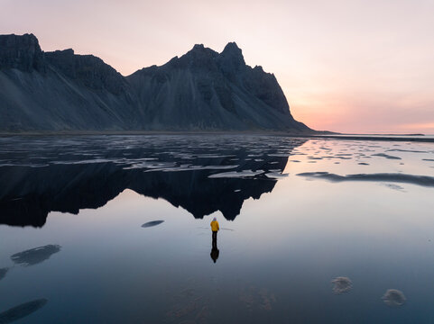 Aerial view of a person in a yellow jacket standing in the shallow reflective waters of Stokksnes with the dramatic Vestrahorn mountain in the background in Iceland.