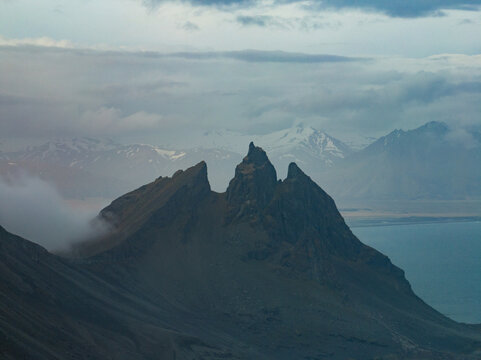 Aerial view of the dramatic peaks of Vestrahorn mountain at Stokksnes, with low-hanging clouds, rugged slopes, and the Atlantic Ocean in the background in Iceland.