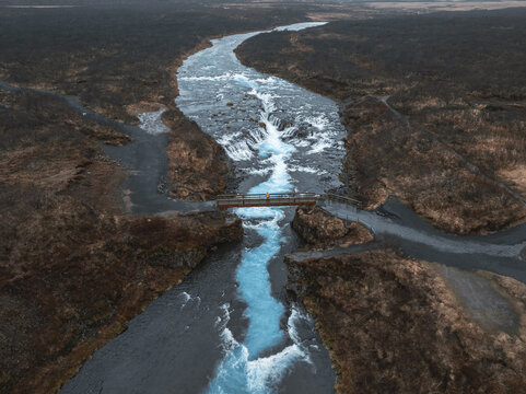 Aerial view of Bruarfoss waterfall with vibrant blue glacial water flowing under a wooden footbridge with a person in a yellow jacket in Blaskogabyggd, Blaskogabyggd, Iceland.