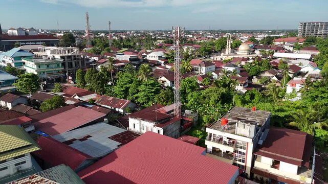 An aerial view showing a BTS (Base Transceiver Station) tower rising above a densely populated tropical urban residential area, with the dome of a mosque visible in the background.