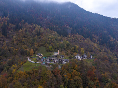 Aerial view of the small village of Camanoglio nestled within a dense forest displaying vibrant autumn foliage on the mountain slopes in Camanoglio, Switzerland.