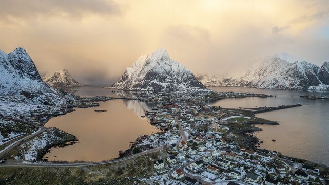 Aerial view of Lofoten, featuring snow-covered mountains, a coastal village with traditional houses, and calm waters reflecting the golden hour light in Nordland, Norway.