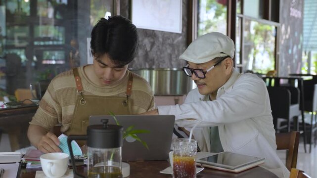 Modern small business digital management young entrepreneur using laptop and tablet planning, finance workflow efficiency technology cafe business operation, learning from senior cafe owner experience