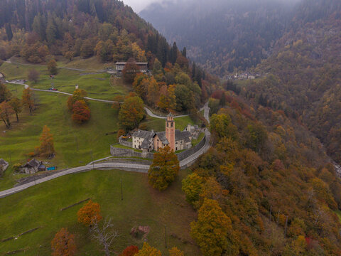 Aerial view of Chiesa di santa Maria surrounded by autumn foliage and winding mountain roads in Chiesa, Cerentino, Switzerland.