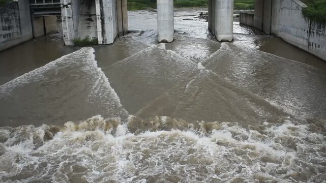 Mud flows beneath concrete bridge supports. Ripples crash against weathered pillars. Floodwaters surge with muddy force. Green shoots cling stubbornly to concrete