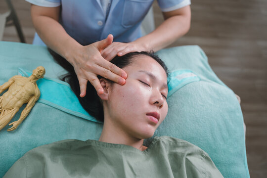Therapeutic facial massage for health wellness concept female therapist hands pressing forehead of young asian woman relieving stress tension near anatomical model in alternative medicine clinic