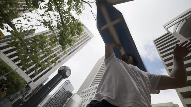 Man Spinning Blue Cross Board Overhead On His Hand - Low Angle With Skyscrapers In San Francisco