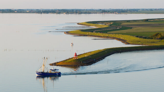 Aerial view of a fishing boat sailing near a lighthouse on a grassy breakwater in the IJsselmeer, with calm water and green coastal fields in Stavoren, Friesland, Netherlands.