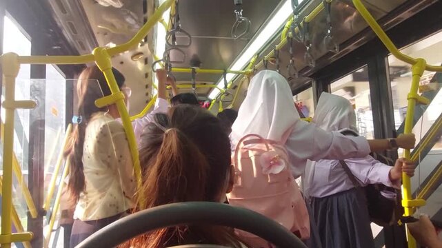 Passengers stand inside a modern city bus during a daily commute, holding handrails while traveling through an urban area with natural daylight entering through the windows.