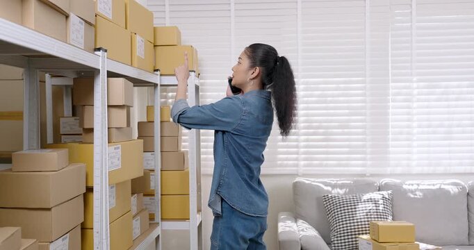 Asian woman small business owner reaches upper shelf while talking with customer on phone discussing product order status and checking warehouse parcel boxes during online seller daily workflow