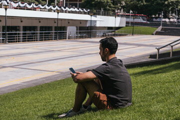 Obraz premium Man sitting on green grass in an urban park, using a smartphone on a sunny day. Digital communication and outdoor leisure