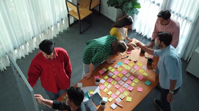 Top view of diverse business people writing and stick sticky notes at whiteboard. Creative team working together and brainstorming marketing idea for startup project at table with notes. Convocation.